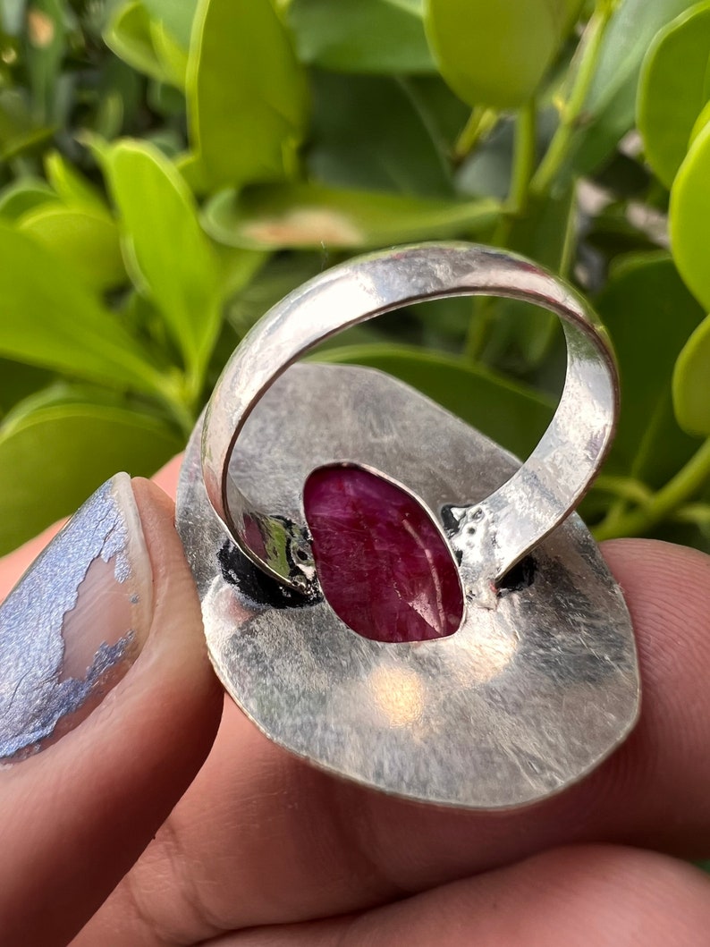 May include: A silver ring featuring a large, oval-shaped, deep pink gemstone set in the centre. The ring is crafted from hammered silver and has a textured finish.