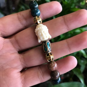 May include: A beaded bracelet with a white Buddha head charm, green jade beads, brown wood beads, and gold accents.