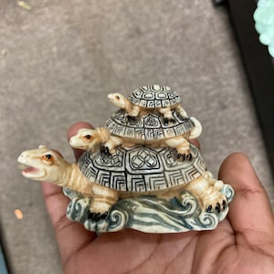 Peut inclure: Une pile de trois petites tortues en céramique blanche avec des marques brunes et noires. Les tortues sont empilées les unes sur les autres, la plus petite étant en haut. Les tortues sont assises sur une base en forme de vague.