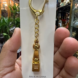 Puede incluir: Un llavero dorado con un pequeño y detallado encanto de torre dorado. El llavero tiene una cadena dorada y un anillo partido dorado. El encanto tiene una base cuadrada con tres líneas horizontales y una punta puntiaguda. El encanto tiene una pequeña bola redonda en la parte superior. El llavero está en una tarjeta blanca con el texto "MADE IN CHINA".