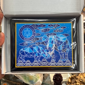 May include: A blue and gold framed print of a geometric elephant and baby elephant on a blue background with white clouds and stars. The print has a circular design with text in a foreign language around the outside.