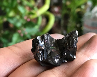 Könnte beinhalten: Ein schwarzer, irisierender Obsidian-Stein mit rauer, zerklüfteter Textur. Der Stein ist etwa so groß wie eine kleine Faust.