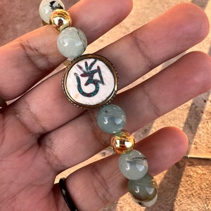 May include: A green and gold beaded bracelet with a circular charm featuring a white Om symbol with turquoise accents.