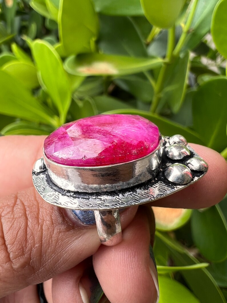 May include: A silver ring featuring a large, oval-shaped pink gemstone set in a bezel setting. The ring has a textured band and a decorative design on the side.