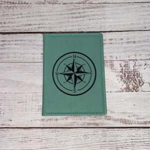 Könnte beinhalten: Ein blaugrüner Reisepass-Etui mit einem schwarzen Kompass-Design. Der Kompass hat die Buchstaben N, S, E und W um den Außenrand.