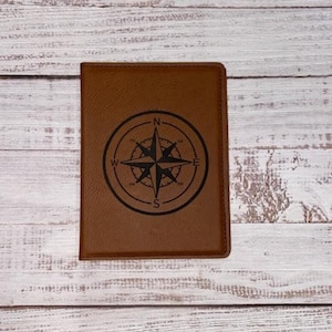Könnte beinhalten: Ein brauner Leder-Reisepasshalter mit einem eingravierten Kompass-Design auf der Vorderseite. Der Kompass zeigt die Himmelsrichtungen N, S, E und W.