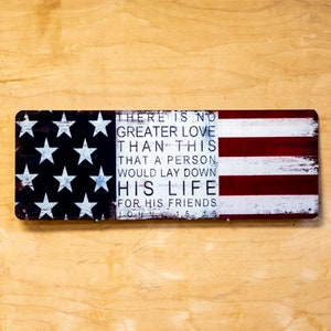 May include: A wooden sign with a distressed American flag design and a quote about love. The quote reads "There is no greater love than this that a person would lay down his life for his friends. John 15:13"