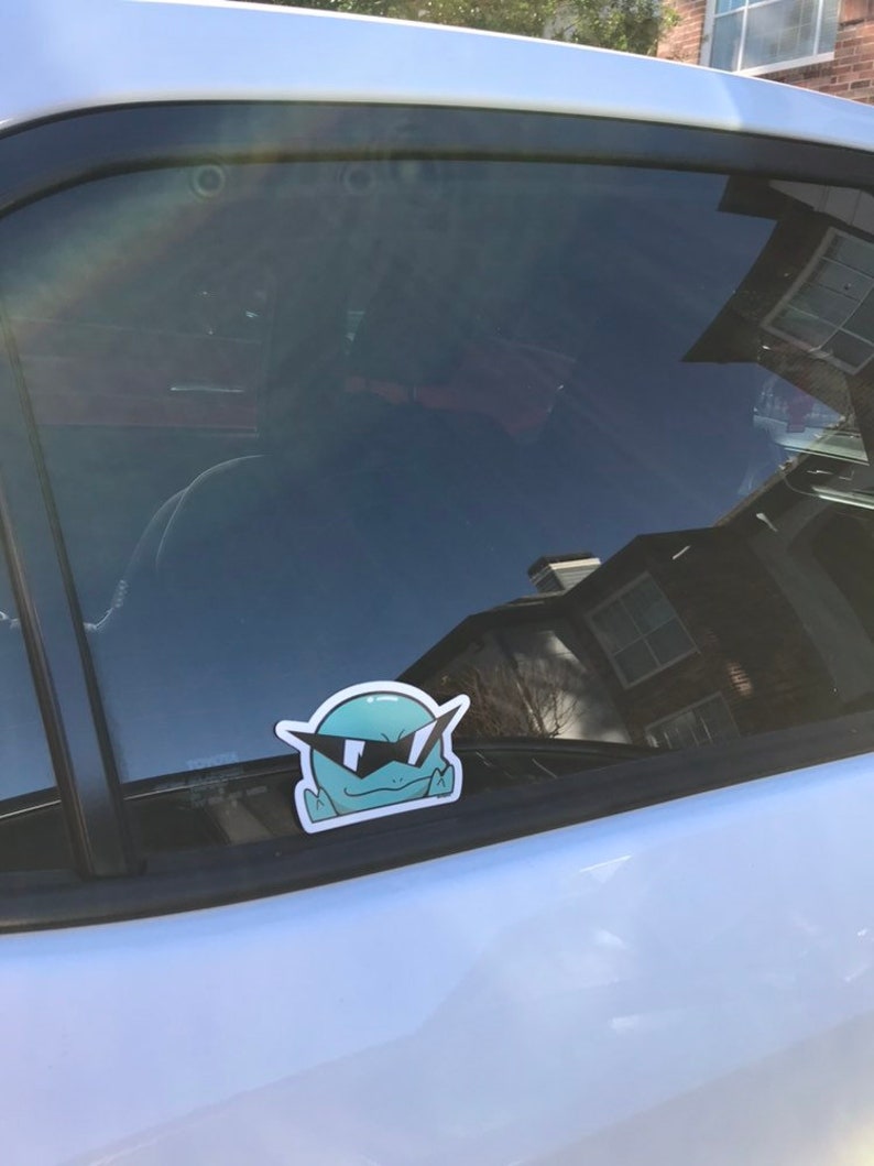 May include: A blue car window with a sticker of a green cartoon character wearing sunglasses. The character has a wide smile and is looking to the right.
