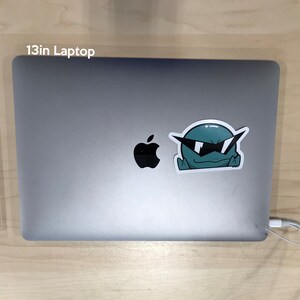 May include: A 13-inch silver laptop with a black Apple logo and a sticker of a green cartoon character wearing sunglasses.