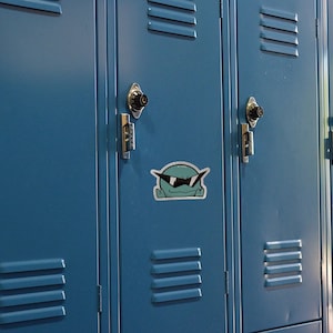 May include: A row of blue metal lockers with a cartoon sticker of a green creature wearing sunglasses on one of the lockers.