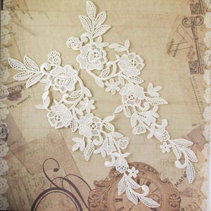 May include: Two off-white lace appliques featuring intricate floral and leaf embroidery. Each piece displays a symmetrical arrangement of roses, small blossoms, and pointed foliage, crafted with detailed stitching and openwork patterns.