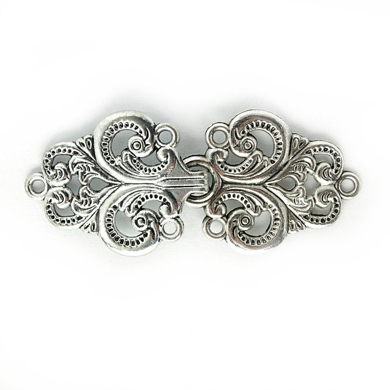 May include: Ornate silver-toned clasp with a symmetrical design. The clasp features intricate scrollwork and decorative details. It has a central locking mechanism and loops on each end for attachment. The clasp is likely used for jewelry or garment closures.