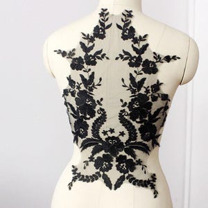 Peut inclure: Une application en dentelle florale noire présentant une broderie complexe sur un support en maille transparente couleur chair. Le motif affiche des dessins botaniques symétriques avec des feuilles et des fleurs détaillées, disposés en forme verticale.