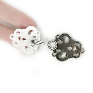 May include: Two antique silver-colored clasps. Each clasp has an ornate, openwork design with a floral motif. One clasp has a cylindrical post, and the other has a ring to receive the post. The clasps are shown against a white background.