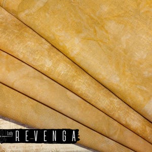 May include: Close-up of folded fabric in shades of yellow and cream. The material has a textured appearance with subtle variations in color, creating a layered effect. The text "Lady Revenga" is displayed in a black rectangle at the bottom.