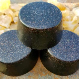 May include: Three cylindrical bars of dark charcoal-colored soap with a subtle copper-toned metallic shimmer throughout the surface. The bars are arranged in a triangular formation with one bar stacked on top of two others, featuring a smooth finish.
