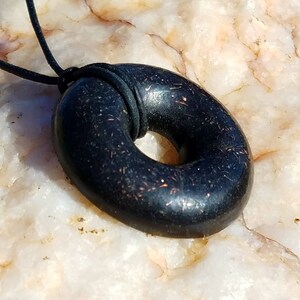 Side angle view of a Black Sun Orgonite donut pendant necklace resting on natural quartz stone. Handmade orgonite jewelry with a polished black surface, rounded edges, and embedded metallic elements. Durable everyday pendant on a black leather cord.