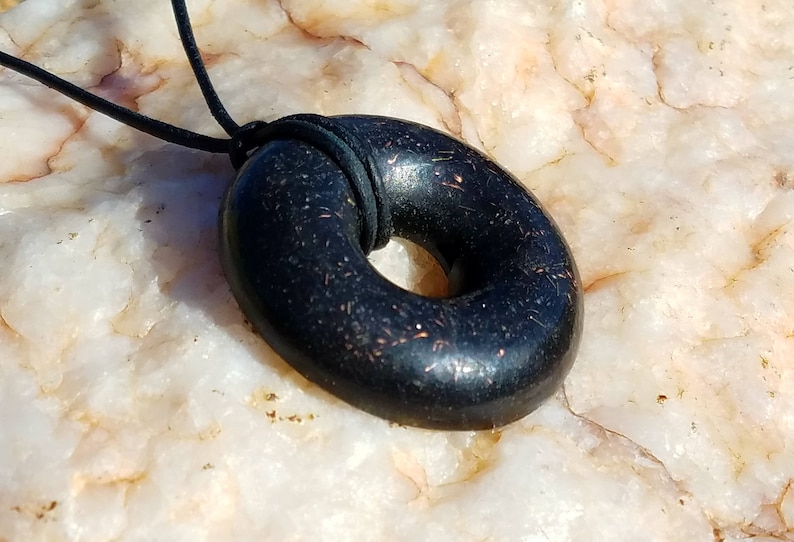 Side angle view of a Black Sun Orgonite donut pendant necklace resting on natural quartz stone. Handmade orgonite jewelry with a polished black surface, rounded edges, and embedded metallic elements. Durable everyday pendant on a black leather cord.