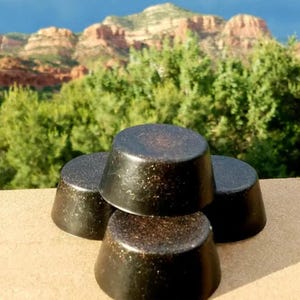 May include: Three round, black, and brown soap bars stacked on top of each other. The soap bars have a rough, textured surface.