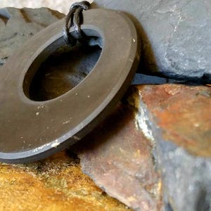 May include: A dark gray, circular pendant with a central hole, suspended by a black cord. The pendant rests against a backdrop of rough, textured rocks in shades of gray, brown, and yellow, creating a natural, earthy aesthetic.