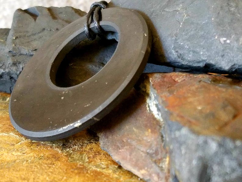 May include: A dark gray, circular pendant with a central hole, suspended by a black cord. The pendant rests against a backdrop of rough, textured rocks in shades of gray, brown, and yellow, creating a natural, earthy aesthetic.