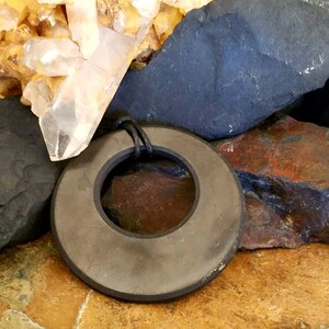 shungite  pendant with a dark gray, matte finish and a black cord. The pendant has a hole in the center and is set against a backdrop of various rocks and crystals, including quartz.