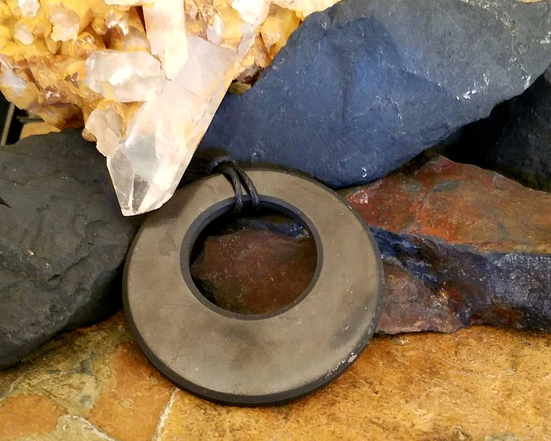 shungite  pendant with a dark gray, matte finish and a black cord. The pendant has a hole in the center and is set against a backdrop of various rocks and crystals, including quartz.