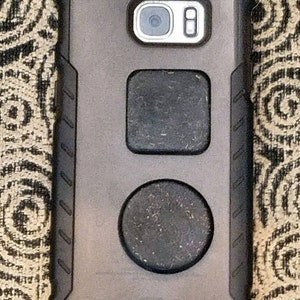 May include: A black phone case with a textured design and two circular cutouts. The cutouts are covered with a black material that appears to be fabric or felt.