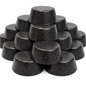 May include: A pyramid-shaped stack of fifteen dark brown, circular, solid bars with a textured, speckled surface. Each individual bar features a flat top and slightly tapered sides, arranged in a symmetrical formation against a white background.