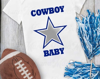 Pode incluir: Um macacão branco para bebê com uma estrela azul e o texto "COWBOY BABY". O macacão é cercado por uma bola de futebol, pompons azuis e brancos e um fundo de madeira.