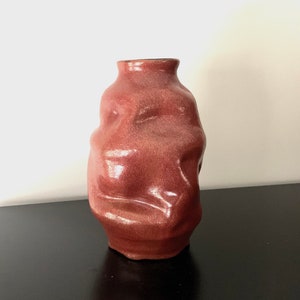 May include: A deep red ceramic vase with an irregular, organic shape. The vase has a smooth, glossy finish.