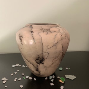 May include: A light pink ceramic vase with a unique black crackle glaze pattern. The vase has a rounded body and a wide opening.