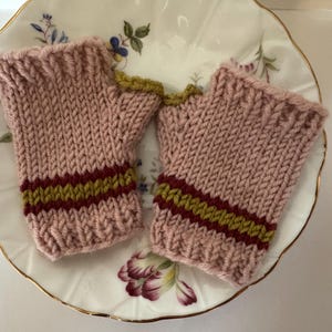 May include: A pair of pink knitted fingerless gloves with a ribbed cuff and a band of burgundy and olive green stripes. The gloves are displayed on a floral patterned plate with a gold rim.