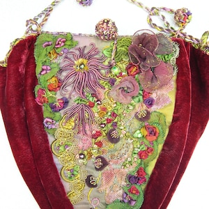 Puede incluir: Una bolsa de terciopelo rojo en forma de corazón con un diseño bordado floral. El bordado presenta una variedad de colores, incluyendo rosa, verde, dorado y morado. La bolsa tiene un cierre de cordón y un borla.