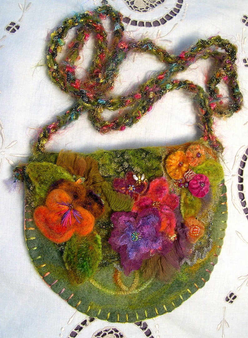 May include: A green felt purse with a floral design. The purse has a long, colourful crocheted strap. The flowers are made of felt and are in shades of orange, red, purple, and green.