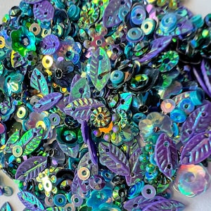 May include: Assortment of iridescent sequins in shades of blue, green, and purple. The sequins are shaped like leaves, flowers, and circles.