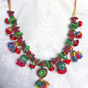 May include: A colourful necklace made of fabric scraps, beads, and tassels. The necklace features a variety of colours, including red, green, blue, and gold. The necklace is strung on a thin cord and has a variety of different shapes and sizes of beads and tassels.