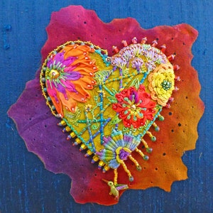 May include: A colourful heart-shaped embroidery featuring a hummingbird design. The embroidery incorporates a range of colours, including red, orange, yellow, green, blue, and purple. The hummingbird is crafted from green, blue, and purple thread. The embroidery is set against a dark blue background.