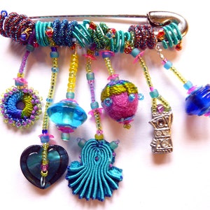 Puede incluir: Un broche colorido con cierre de pasador de seguridad y colgantes. Los colgantes incluyen un corazón, una cuenta de vidrio azul, una bola de tela rosa y verde, un círculo de tela azul y verde, y un amuleto plateado con pedrería. El broche está hecho con una variedad de cuentas, tela y alambre.