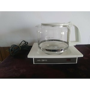 May include: A white Mr. Coffee coffee maker with a glass carafe. The carafe has a white lid and a black handle. The coffee maker has a black base with a red light on the front.
