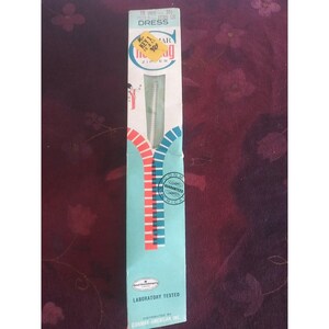 May include: A white zipper packaged in a blue and red box. The box has the text "Conmar Key 1 Zipper" and "Laboratory Tested" printed on it. The zipper is 25.4 cm long.