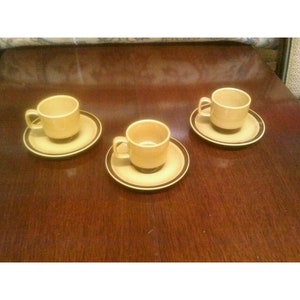 以下が含まれることがあります： 茶色とベージュのセラミック製のコーヒーカップ3つと、それに合う受け皿。カップと受け皿には、縁に茶色の帯がある。
