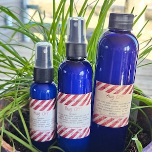 May include: Three blue bottles of bug spray with red and white striped labels. The labels read "Bug Off!" and list ingredients. The bottles are arranged in order of size, with the largest bottle on the right and the smallest bottle on the left.