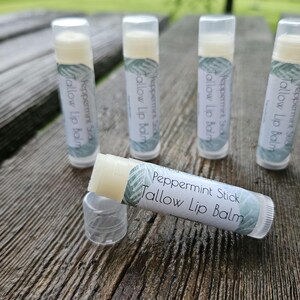 May include: Several tubes of peppermint stick tallow lip balm are arranged on a wooden surface. The tubes are white with labels that read "Peppermint Stick Tallow Lip Balm" in a script font. The lip balm is a light yellow color.