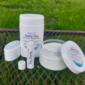 May include: Three white containers of sunscreen products with the text "simply good Sunny Day Natural Water Resistant Sunscreen" on the labels. The containers are on a metal surface.