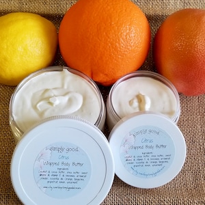 May include: Two jars of white whipped body butter with a citrus scent. The jars have white lids with black text that reads "simply good. Citrus Whipped Body Butter" and lists the ingredients. The jars are sitting on a brown burlap surface with three citrus fruits: a lemon, an orange, and a grapefruit.
