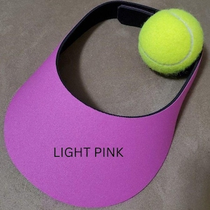 May include: A light pink visor with a black band and a yellow tennis ball. The text "LIGHT PINK" is printed on the visor. The visor is designed to shield the eyes from the sun.