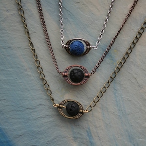 May include: Three layered necklaces with lava stone beads in a cage setting. The top necklace has a blue stone, the middle necklace has a black stone, and the bottom necklace has a black stone. The necklaces are on a light blue background.