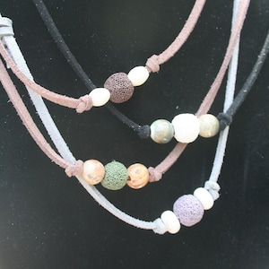 May include: A selection of layered necklaces with suede cords in black, grey, and brown. Each necklace features a variety of beads, including white, brown, green, and purple, creating a natural aesthetic. The necklaces are displayed on a black mannequin.