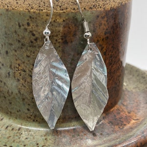 May include: A pair of silver leaf earrings with a textured finish. The earrings are hanging from a brown ceramic mug.